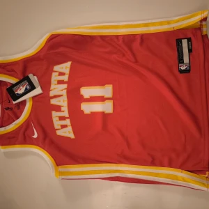 NBA JERSEY - ATLANTA - Snygg röd basketlinne från Nike med gula detaljer och texten 'ATLANTA' samt nummer 11 på framsidan. Perfekt för basketfans och sportentusiaster. Officiell NBA Authentic Icon Edition.
