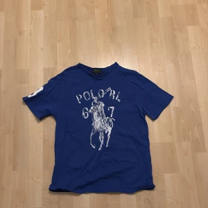 Blå t-shirt från Polo Ralph Lauren - Säljer en blå t-shirt från Polo Ralph Lauren med ett stort tryck av en polospelare och texten 'Polo RL 67' på framsidan. T-shirten har korta ärmar och är tillverkad i bomull. Perfekt för en avslappnad stil. Tröjan är i storlek m (10-12)!