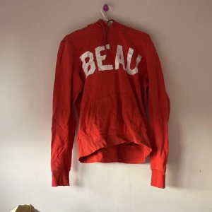 Röd hoodie med text - Snygg röd hoodie med stor vit text 'BEAU' på framsidan. Tröjan har en bekväm passform med långa ärmar och en praktisk huva. Perfekt för en avslappnad stil.