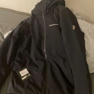 Svart jacka från Peak Performance - Säljer en stilren svart jacka från Peak Performance med huva och dragkedja. Perfekt för utomhusaktiviteter med sin funktionella design. Aldrig använd lappen sitter kvar som ni ser på den, ny pris 3500 men kan sälja vid 1000 vid snabb affär 