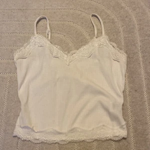 Vit ribbad linnetopp med spetsdetaljer - Säljer en söt vit ribbad linnetopp med smala axelband och spetsdetaljer längs halsringningen och nederkanten. Perfekt för sommardagar eller som ett lager under en tröja.