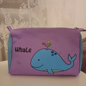 Väska - Söt lila necessär med en blå val och texten 'Whale' på framsidan. Väskan har en dragkedja upptill och är perfekt för att förvara småsaker. Den har ljusblå sidor som ger en fin kontrast.