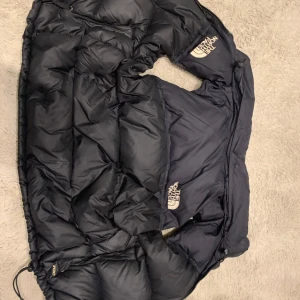 Svart dunjacka från The North Face - Säljer en svart dunjacka från The North Face. Jackan har en pufferdesign med dragkedja och är perfekt för kalla dagar. Den är fylld med äkta dun för extra värme och komfort.