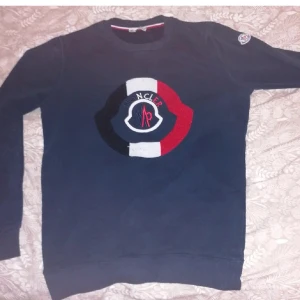 Mörkblå sweatshirt från Moncler - Snygg mörkblå sweatshirt från Moncler med ett stort, färgglatt logotypmärke på bröstet i rött, vitt och blått. Tröjan har långa ärmar och rund halsringning. Perfekt för en stilren och avslappnad look.