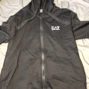 Armani hoddie - Säljer en stilren svart hoodie från Emporio Armani EA7 med dragkedja och huva. Perfekt för en casual look med sitt enkla men eleganta design. Märkesloggan är tryckt på bröstet. Size M