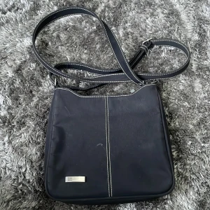 Svart axelväska från Zera - Snygg svart axelväska från Zera med justerbar axelrem och stilren design. Väskan har en metallplatta med logotyp på framsidan och kontrastsömmar som ger en extra touch. Perfekt för att bära dina dagliga nödvändigheter med stil.