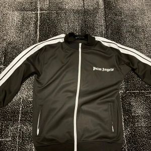 Palm Angels kofta - Snygg svart träningsjacka från Palm Angels med vita ränder längs ärmarna och dragkedja framtill. Jackan har en sportig look med ribbade muddar och två fickor med dragkedja.