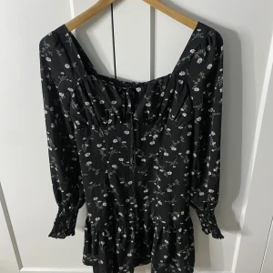 Svart blommig klänning från Boohoo - Charmig svart klänning från Boohoo med ett blommigt mönster. Klänningen har långa ärmar och en volangdetalj vid fållen. Perfekt för en stilfull look. Den jag även en buschering vid bysten.