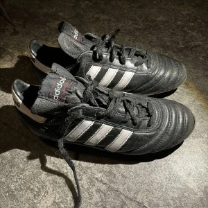 Svarta fotbollsskor från Adidas - Klassiska svarta fotbollsskor från Adidas med vita ränder och snörning. Perfekta för gräsplaner med sina dobbar. Skorna har en tidlös design och är idealiska för fotbollsspelare som söker både stil och funktion.