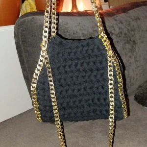 Svart virkad väska med guldkedjor - Trendig svart virkad väska med iögonfallande guldkedjor. Perfekt för att ge din outfit en unik touch. Väskan har en mjuk och flexibel design.