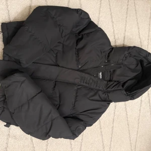 Svart pufferjacka från Trapstar - Snygg svart pufferjacka från Trapstar med huva och dragkedja. Jackan har en broderad logga på ryggen och är perfekt för kyligare väder. Ny pris 4500! Använd ett par gånger ny skick