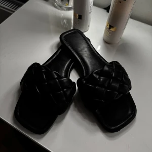 Svarta flätade sandaler - Snygga svarta sandaler med flätad design. Perfekta för en stilren look. De har en platt sula och är tillverkade i syntetmaterial. Liten skada framtill!