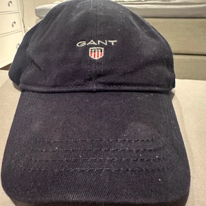 Mörkblå keps från GANT - Snygg mörkblå keps från GANT med broderad logotyp framtill. Kepsen har en justerbar rem baktill för perfekt passform. Perfekt för en avslappnad stil.