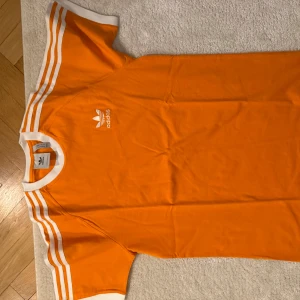 Orange t-shirt från Adidas - Snygg orange t-shirt från Adidas med klassiska vita ränder på ärmarna och logga på bröstet. Perfekt för en sportig look eller avslappnad stil. Kortärmad och bekväm passform. Storlek M. Mycket bra skick.