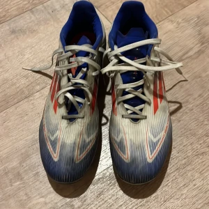 Adidas fotbollsskor i vitt, blått och rött - Säljer ett par Adidas fotbollsskor i vitt med blå och röda detaljer. Skorna har snörning och en sportig design, perfekt för fotbollsplanen. De är tillverkade i syntetmaterial och har en bekväm passform. Priset kan diskuteras!