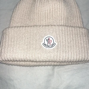 Beige stickad mössa från Moncler - Snygg beige stickad mössa från Moncler med deras ikoniska logga framtill. Perfekt för att hålla dig varm och stilren under kyliga dagar.