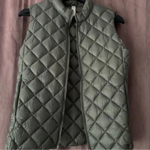 Grön quiltad väst - Snygg grön quiltad väst med dragkedja framtill. Perfekt för lager-på-lager under kyligare dagar. Västen har en stilren design med ett klassiskt rutmönster.