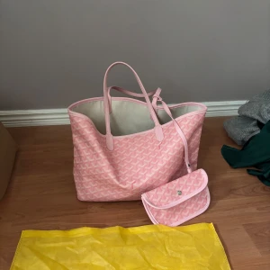 Rosa handväska  - Säljer en snygg rosa goyardliknande väska med matchande mönster och en liten plånbok. Väskan har två handtag och en praktisk innerficka. Perfekt för att bära med sig allt man behöver. Köpt utomlands för 1500. En fläck i botten av en penna men inget som syns utåt. 