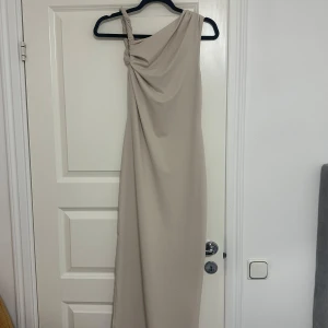 Beige axelbandslös långklänning - Elegant beige långklänning med en unik axelbandslös design och en tvinnad axelrem. Klänningen har en slits som ger en stilfull touch. Perfekt för speciella tillfällen.