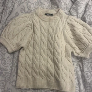 Vit stickad tröja från Gina Tricot - Säljer en söt vit stickad tröja från Gina Tricot med korta puffärmar och ett klassiskt flätmönster. Perfekt för en stilren look. Tröjan har ribbade kanter vid ärmslut och nederkant. Passar bra till både jeans och kjol.