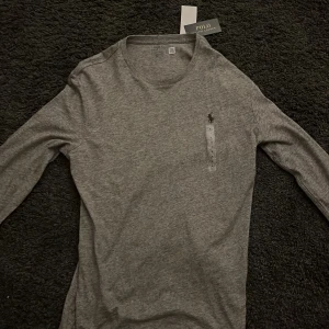 Grå långärmad tröja från Ralph Lauren - Säljer en stilren grå långärmad tröja från Ralph Lauren. Tröjan har en broderad logotyp på bröstet och är tillverkad i mjukt material för extra komfort. Köpt i en Ralph Laurent butik i usa och inte använd en enda gång sedan dess. Den är i storlek m.