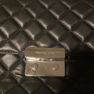 Svart quiltad axelväska från Michael Kors - Snygg svart axelväska från Michael Kors i quiltat skinn. Väskan har en elegant kedjerem och ett stilrent metallspänne framtill. Perfekt för att ge din outfit en lyxig touch.