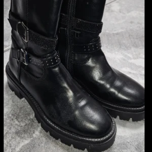 Svarta boots med spännen - Snygga svarta boots i skinn med dekorativa spännen och nitar. Perfekta för en tuff look med robust sula och dragkedja på sidan för enkel på- och avtagning.