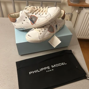 Vita sneakers från Philippe Model - Snygga vita sneakers från Philippe Model med unika detaljer i blått och brunt. Skorna har snörning och en klassisk design som passar perfekt för en stilren look. Levereras med originalpåse. Ordinarie pris ligger på 3800kr och dom är helt oanvända.  
