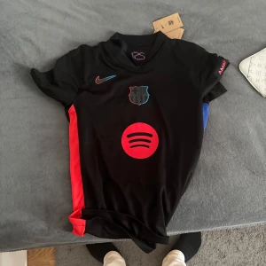 Svart fotbollströja med röda detaljer - Snygg svart fotbollströja från Nike med röda och blå detaljer. Tröjan har korta ärmar och ett tryck med nummer 19 och namnet Lamine Yamal på baksidan. Perfekt för fotbollsfans som vill visa sitt stöd.