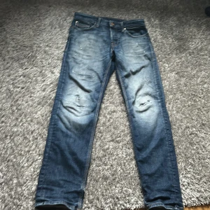 Blå jeans med slitningar - Säljer ett par blå jeans med coola slitningar och en klassisk femficksdesign. De har en tapered passform och en snygg läderdetalj på baksidan. Perfekta för en avslappnad stil.