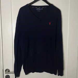 Mörkblå tröja från Ralph Lauren - Säljer en stilren mörkblå tröja från Ralph Lauren i Pima bomull. Tröjan har en v-ringad design med ett rött broderat märke på bröstet. Perfekt för en klassisk och avslappnad look. Färg marinblå. Skick 9/10. Skriv om ni har några frågor.