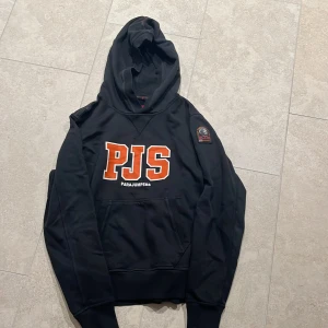 Svart hoodie från Parajumpers - Säljer en svart hoodie från Parajumpers med stor PJS-logga i orange och vitt på framsidan. Hoodien har en klassisk känguruficka och en patch på ärmen. Tillverkad i mjukt fleecematerial för extra komfort. 