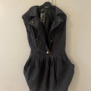 Mörk klänning med dragkedja - Snygg mörk klänning med dragkedja framtill och krage. Klänningen har en avslappnad passform med fickor och är ärmlös, vilket gör den perfekt för varmare dagar.