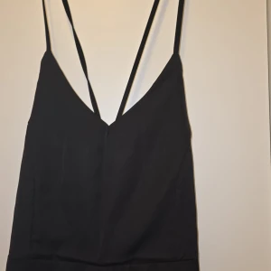 Svart långklänning med snörning - Elegant svart långklänning med smala axelband och djup v-ringning. Klänningen har en stilren design med snörning i ryggen för en justerbar passform. Perfekt för en kväll ute eller en speciell tillställning.