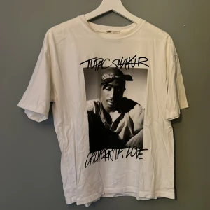 Vit T-shirt med Tupac-tryck från Pull&Bear - Säljer en vit T-shirt från Pull&Bear med ett stort tryck av Tupac Shakur på framsidan. T-shirten har korta ärmar och en rund halsringning. Perfekt för dig som älskar klassisk hiphop-stil. 🖤