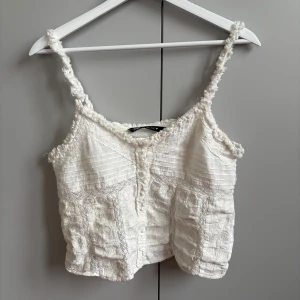 Vit linnetopp från Zara - Säljer en söt vit linnetopp från Zara med tunna axelband och dekorativa sömmar. Toppen har en lätt textur och är perfekt för varma dagar. Den är både stilren och bekväm.