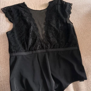 Svart spetsbody - Svart body med spetsdetaljer och djup v-ringning baktill och framtill, men med Mesh fram. Den har korta ärmar och är i string-modell. 