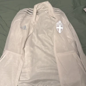 Vit vindjacka från Adidas - Säljer en vit kofta från Adidas med Sveriges emblem på bröstet och texten 'Sverige' på ryggen. Jackan har dragkedja och långa ärmar med de klassiska Adidas-ränderna på axlarna. Perfekt för sportiga tillfällen!
