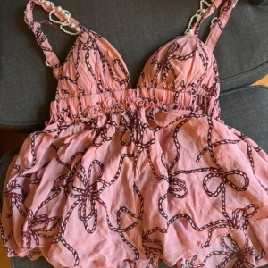 Rosa sommartopp från Pinko - Säljer en söt rosa topp från Pinko med ett unikt mönster av svarta rep och pärldetaljer på axelbanden. Klänningen har en v-ringad design och elastisk midja för en bekväm passform. Perfekt för sommarens äventyr! 🌸