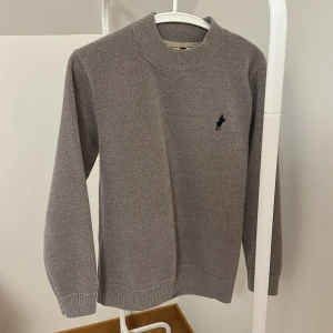 Grå tröja från Ralph Lauren - Säljer en stilren grå tröja från Ralph Lauren med deras ikoniska logga broderad på bröstet. Tröjan har långa ärmar och en rund halsringning, perfekt för en avslappnad men ändå elegant look.