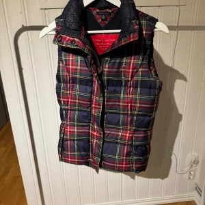Rutig dunväst från Tommy Hilfiger - Snygg rutig dunväst från Tommy Hilfiger i blått, rött och gult. Västen har dragkedja och knappar framtill samt en hög krage för extra värme. Perfekt för kyligare dagar.