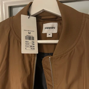 Beige bomberjacka från Kappahl - Säljer en snygg beige bomberjacka från Kappahl i storlek M. Jackan har en dragkedja framtill och ribbade muddar vid ärmslut och nederkant. Perfekt för en stilren look. Färg: Brons.
