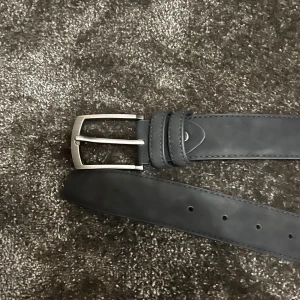 Svart bälte i skinn - Säljer ett stilrent svart bälte i skinn och mocka med en klassisk silverfärgad spänne. Perfekt för både vardag och fest. Bältet har flera hål för justerbar passform och är i nyskick. Passar till alla outfits och ger en snygg touch till din stil.