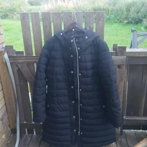 Svart dunjacka från Lindex - Säljer en svart dunjacka från Lindex i storlek XL. Jackan är perfekt för kalla dagar med sin långa design och quiltade mönster. Den har en dragkedja och knappar framtill för extra skydd mot kylan. Huvan ger extra värme och skydd mot väder och vind. Perfekt för vintern!