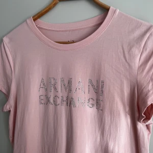 Rosa t-shirt från Armani Exchange - Säljer en superfin rosa t-shirt från Armani Exchange. Den är kortärmad och har ett glittrigt tryck med märkesnamnet på framsidan. 