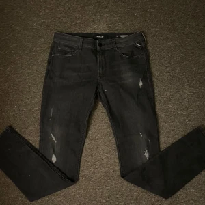 Svarta jeansbyxor från Replay - Snygga svarta jeansbyxor från Replay med slitna detaljer. De har en klassisk femficksdesign och en knappgylf. Perfekta för en avslappnad stil.