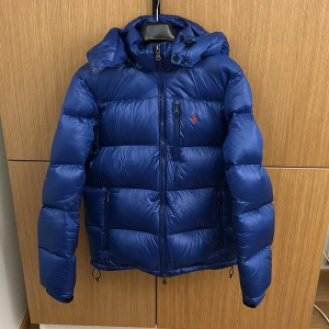 Blå dunjacka från Polo Ralph Lauren - Säljer en snygg blå dunjacka från Polo Ralph Lauren. 9/10 skick syns knappt att den är använd förutom hängaren som lossnat(bild5). Den är perfekt för kyliga dagar under hela vintern. Jackan är både stilren och funktionell. Ställ frågor pris kan diskuteras!