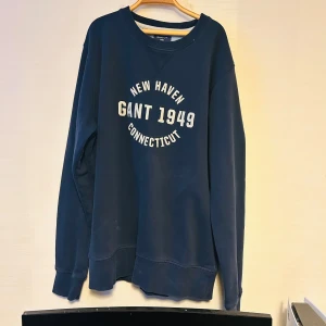 Mörkblå sweatshirt från Gant - Säljer en snygg mörkblå sweatshirt från Gant med tryck 'New Haven Gant 1949 Connecticut' på framsidan. Tröjan har långa ärmar och en rund halsringning. Perfekt för en avslappnad stil.