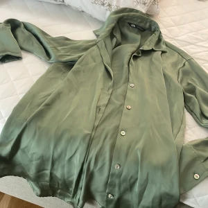 Grön skjorta från Zara - Snygg grön skjorta från Zara i ett mjukt och glansigt material. Skjortan har långa ärmar och knappar framtill. Perfekt för en stilren look. Storlek passar till M 
