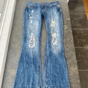 Blå bootcut jeans med slitningar - Snygga blå bootcut jeans med slitningar och unika detaljer på bakfickorna som jag köpt på Plick. Jeansen har en klassisk femficksdesign och en dekorativ lapp med blommönster baktill. Perfekta för en avslappnad stil.